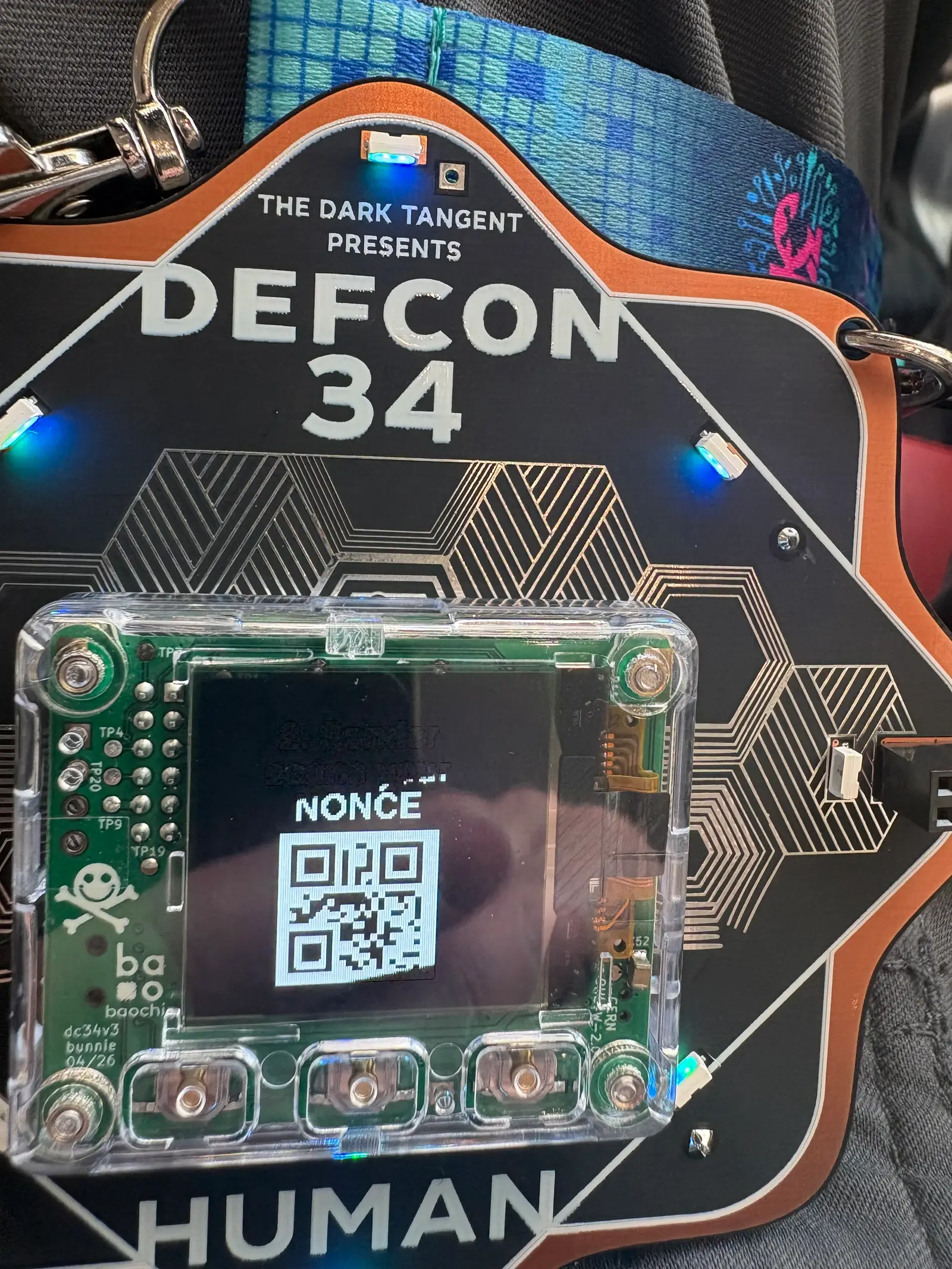 The DEF CON 34 human badge, front on, its screen showing a QR code. Blue LEDs around the outer edge are lit.